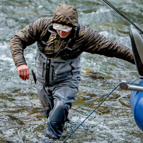An angler wearing zip-front waders steadies themself against their raft as they wade in turbulent water